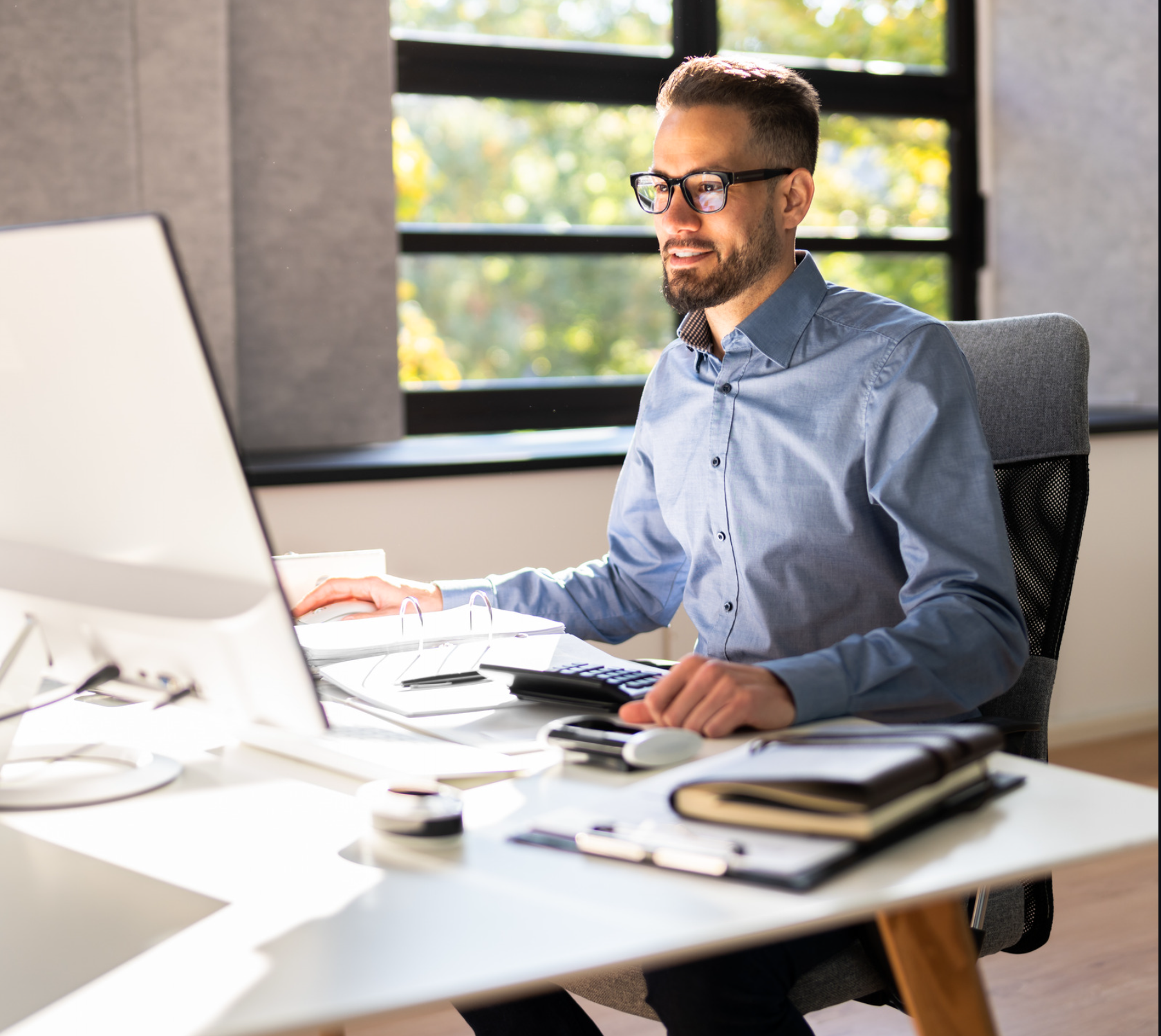 Mann mit Brille arbeitet am Schreibtisch mit Monitor, Taschenrechner und Unterlagen